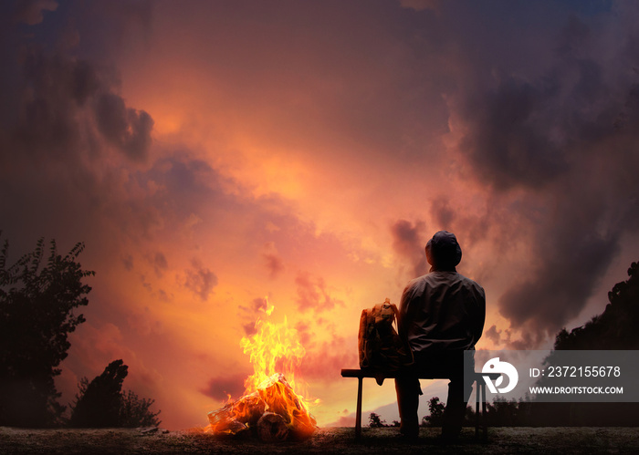 日落时，男性旅行者坐在篝火旁的长椅上。