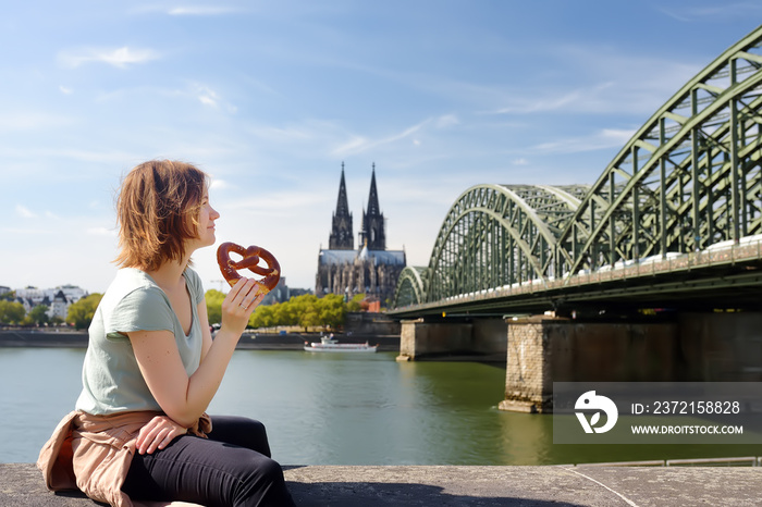 Young woman eats traditional pretzel sitting on embankment of Rhine on background of Cologne Cathedr