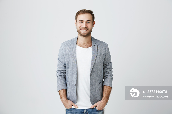 Delightful european man wearing jacket over white t-shirt and jeans, looks pleasantly at camera, has