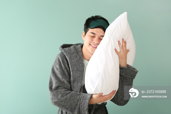 Handsome young man with sleep mask and pillow on color background