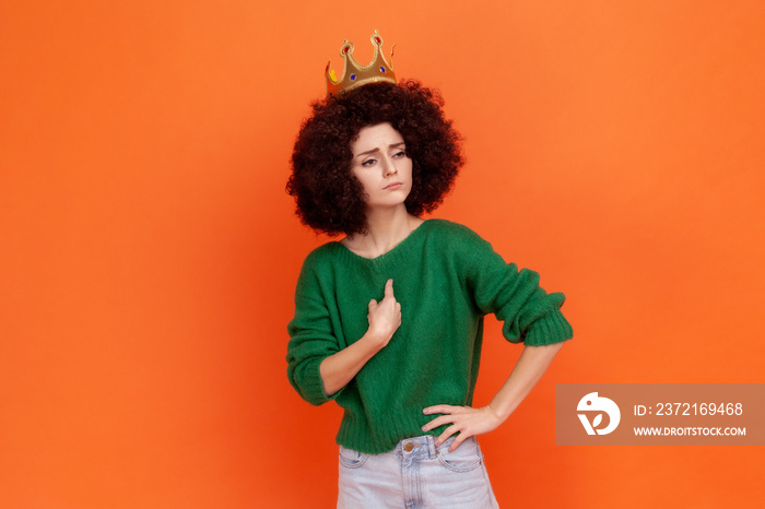 Proud woman with Afro hairstyle wearing green casual style sweater and crown pointing at herself, ha