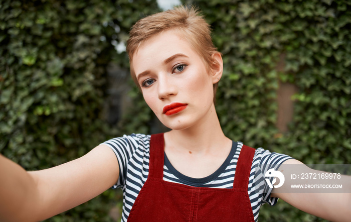 portrait of a blonde with a short haircut bright makeup Red sundress and green leaves of bushes in t