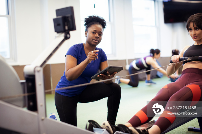 Portrait of instructor using tablet computer while guiding athlete exercising on rowing machine in g