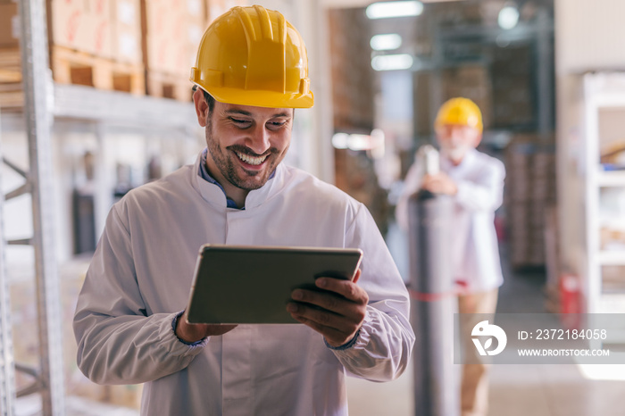 Close up of young Caucasian smiling worker with helmet on head using tablet for work while standing 