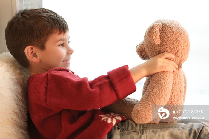 Cute little boy with teddy bear near window at home