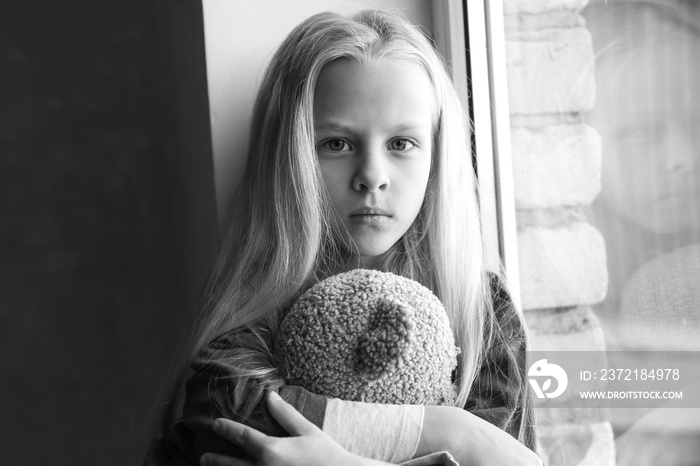 Homeless little girl with teddy bear sitting on window sill