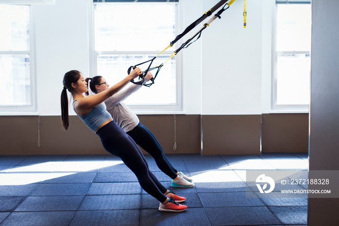 Side view of female athletes pulling suspension straps against windows in gym