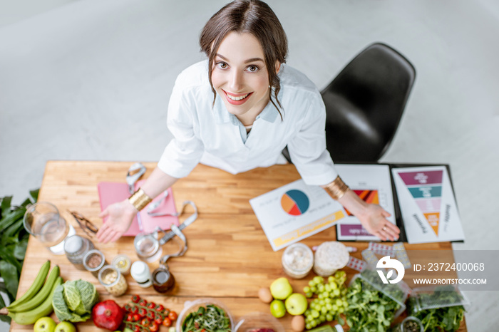 Portrait of a young woman nutritionist in medical uniform standing near the table full of various he