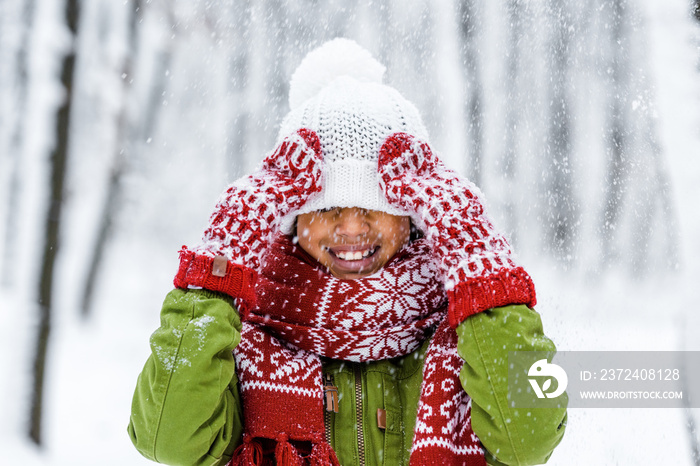 smiling african american child with knitted hat pulled over eyes during snowfall in winter park