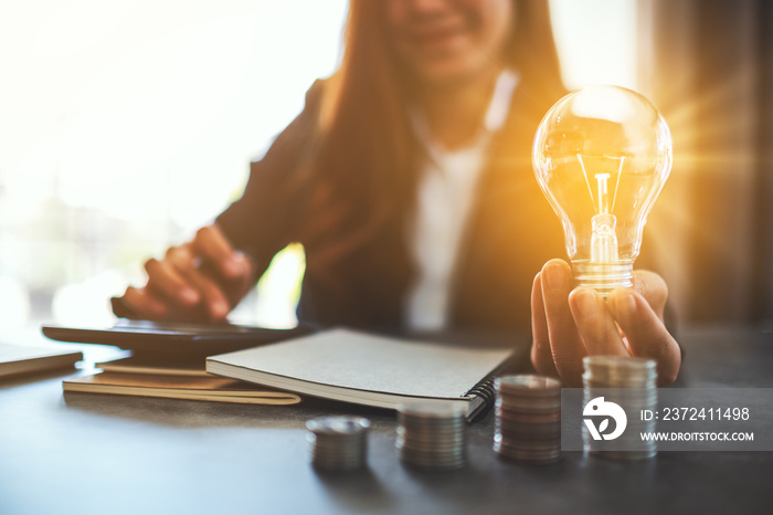 Businesswoman holding a lightbulb with coins stack on table, saving energy and money concept