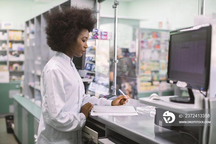 Portrait of young professional concentrated African female pharmacist working with computer behind c