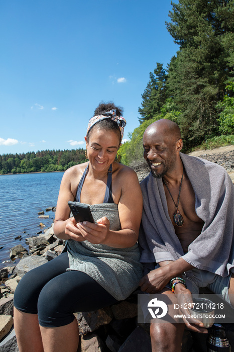 Smiling couple in towels looking at smart phone on lakeshore, Yorkshire, UK