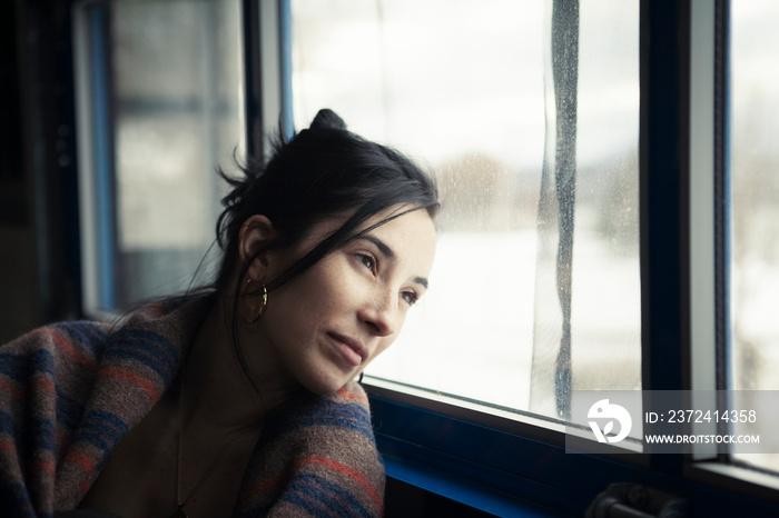 Thoughtful woman wrapped in blanket looking through window while sitting at home