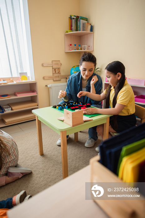 asian girl playing wooden cubes game with teacher near kids on floor in classroom