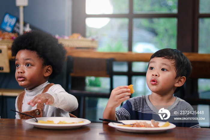 Cute African American boy with curly and adorable Asian kid eating meal at the table indoor, happy c