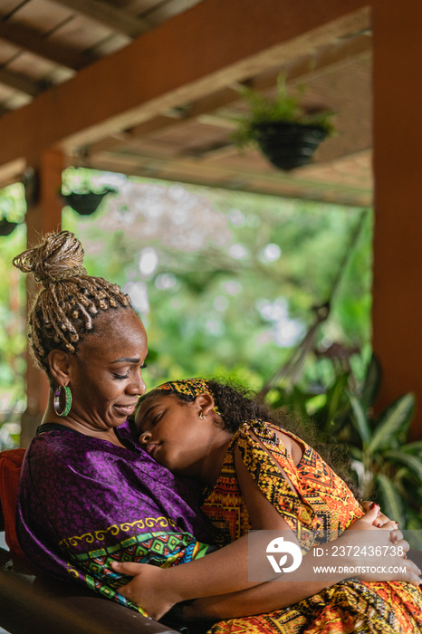 Imagen vertical de una madre afrocaribeña sentada cargando a su hija con mucho amor mientras duerme.