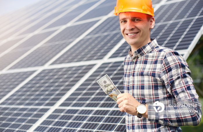 Smiling male with hundred dollar bill in hand at the background of solar panels. Business and genera