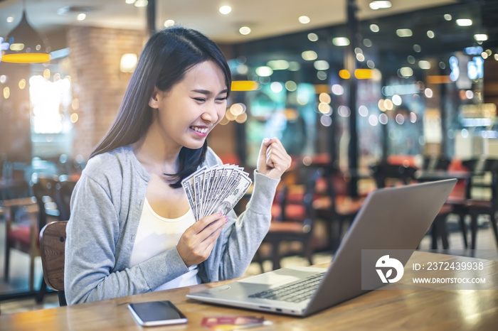 Young woman holding a bunch of dollar banknotes while working online with her laptop. Make money wit