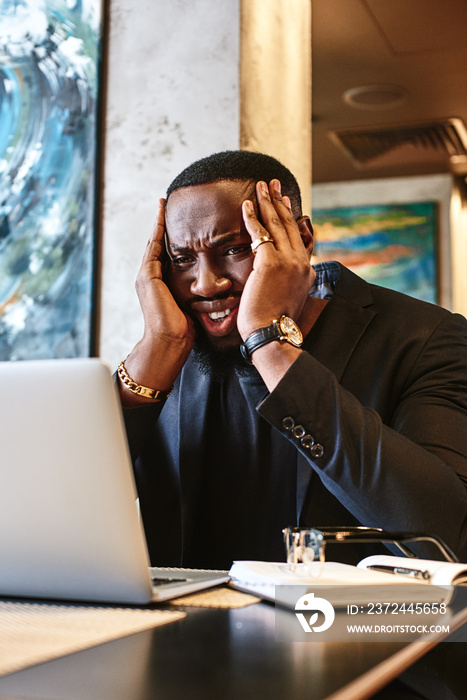 Designer worry about deadline of new project. Man hold head by two hands and look in laptop with pen