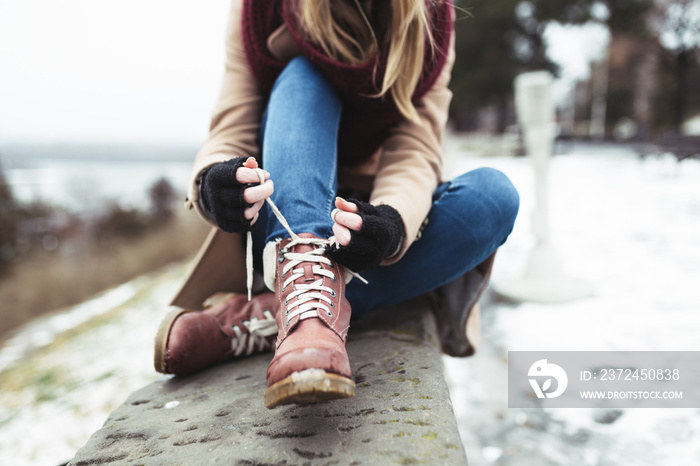 Close up shot of fashionable young woman sitting on stone wall and tying up laces on her boots.