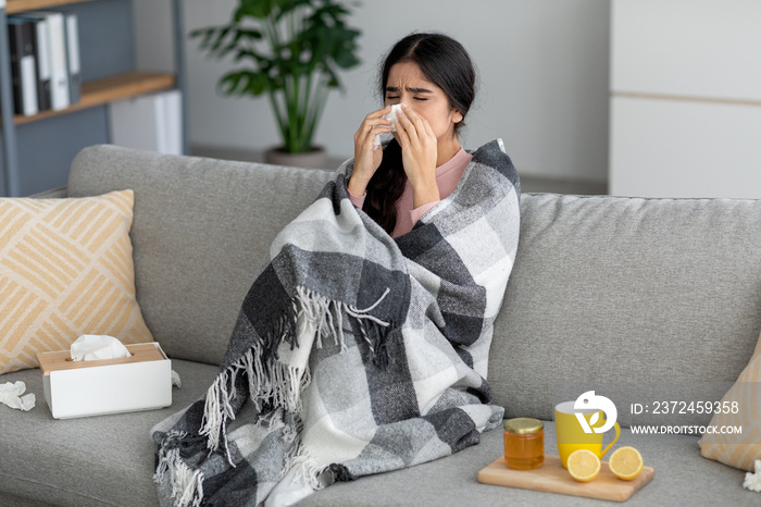 Unhappy young indian lady sneezes in napkin, blows nose, suffers from fever, flu on couch in room in