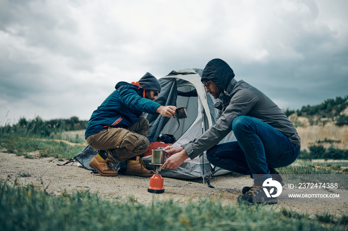 雨天，两名男子蹲在帐篷附近准备咖啡。露营旅行概念。