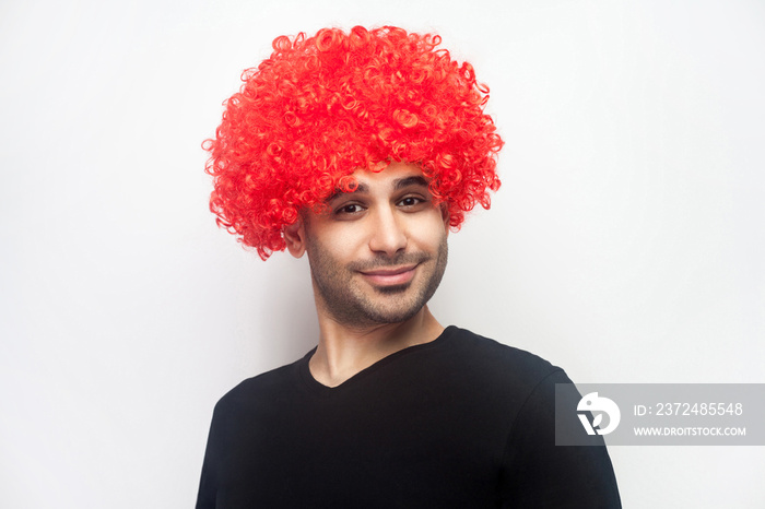 Portrait of funny stylish hipster man with bristle and curly red wig on his head smiling at camera, 
