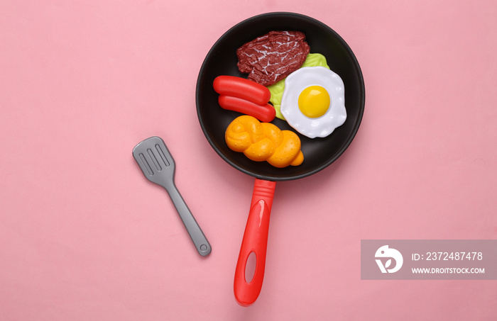 Toy frying pan with scrambled eggs, meat, sausage, bun on pink background with fork