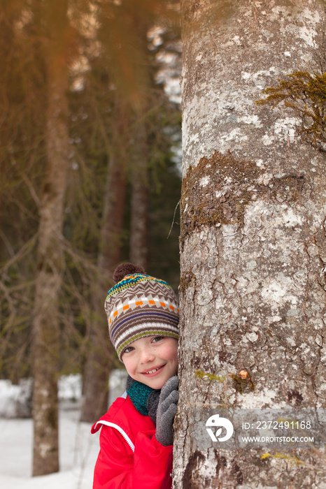 Portrait playful boy hiding behind tree in snowy woods