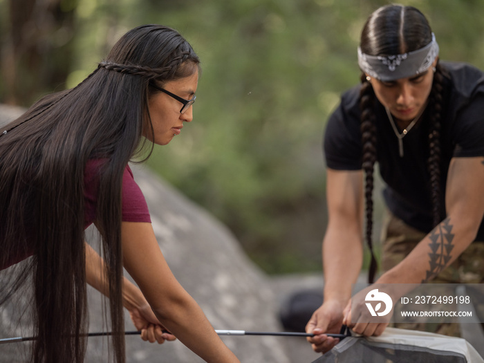 Young native man and native woman helping each other slide tent poles into a tent in the outdoors