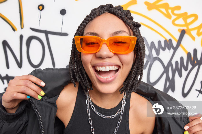 Swag. Fashionable cheerful hipster girl with braids smiles broadly shows white teeth wears trendy or