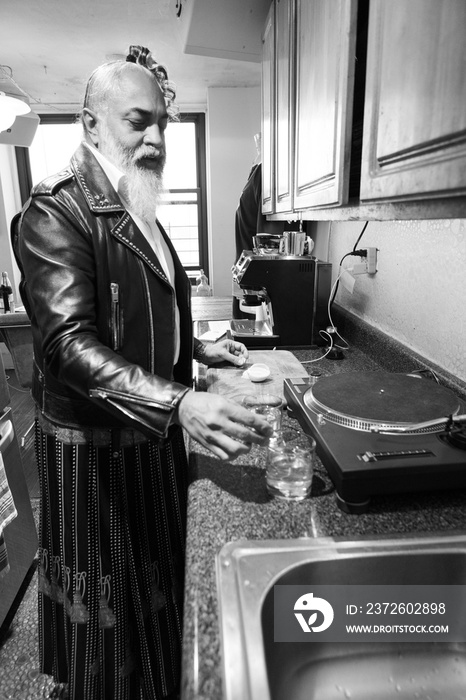 gray haired bearded man in leather jacket prepares a drink in kitchen