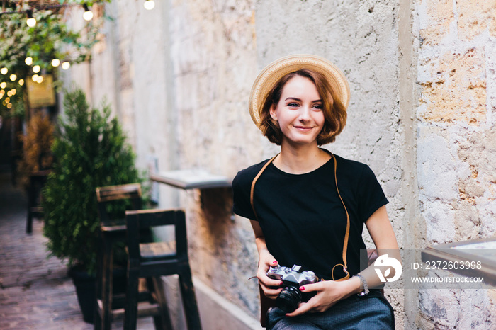 Young hipster with retro camera in hands sits on chair and smiles outdoors against the backdrop of g