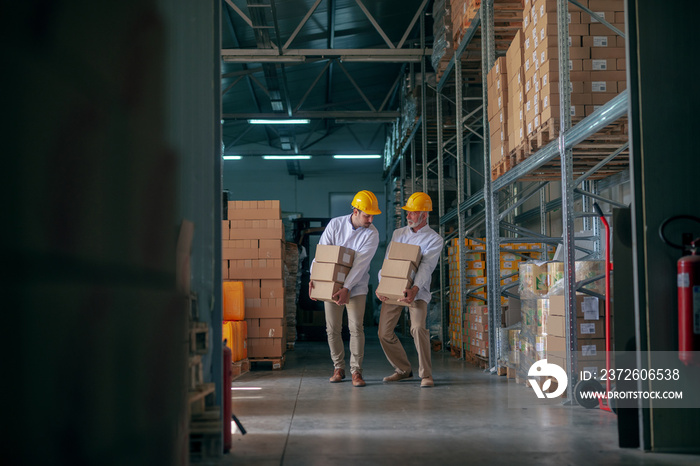 Two storage workers dressed in white uniform and with yellow helmets carrying heavy boxes.