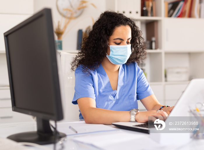 Young woman doctor assistant in protective face mask working in medical office using laptop computer