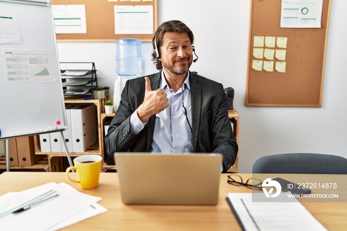 Handsome middle age man wearing call center agent headset at the office doing happy thumbs up gestur