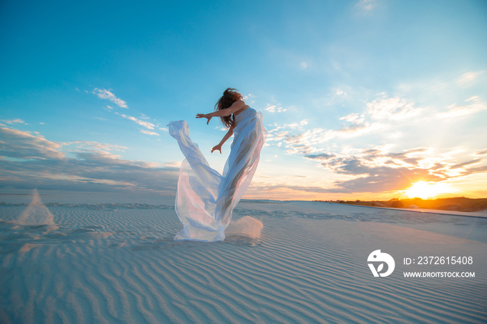 A girl in a fly white dress dances and poses in the sand desert at sunset