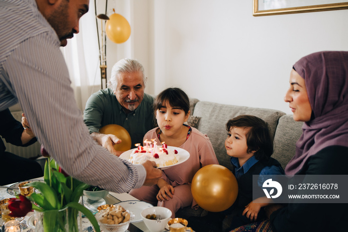 Girl blowing out candles on cake while sitting with family during birthday party