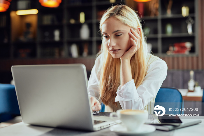 Young serious Caucasian blonde businesswoman dressed smart casual sitting in cafe and using laptop.