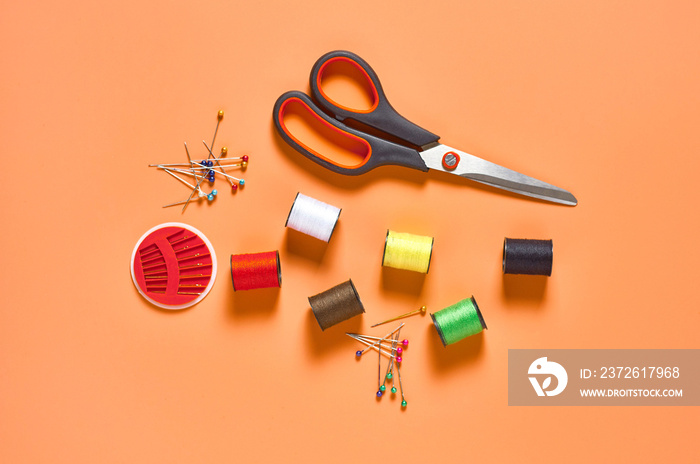 Scattered sewing accessories needles, scissors and spools with colorful threads on orange tailors de