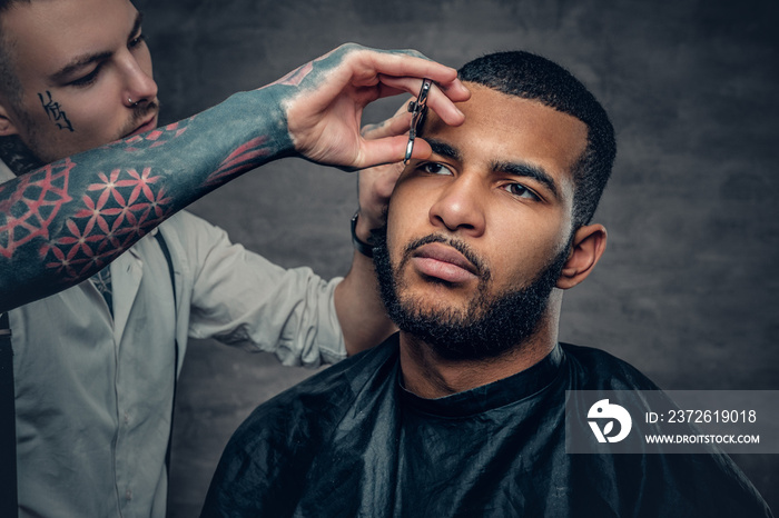 Tattooed barber makes haircut to a Black bearded male.