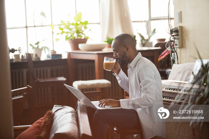 Side view of man having coffee using laptop computer while sitting on stool against window at home