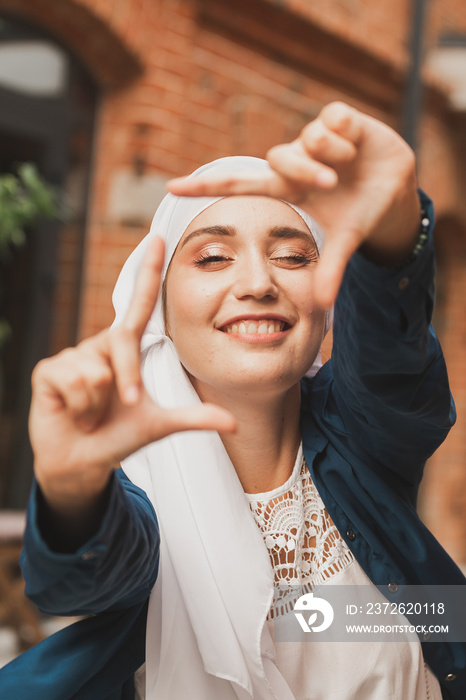 Portrait of young muslim girl making a camera frame with fingers outdoors.