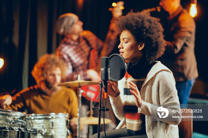 Mixed race woman singing. In background band playing instruments. Home studio interior.