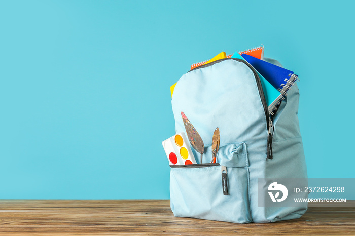 Backpack with school supplies on wooden table against color background