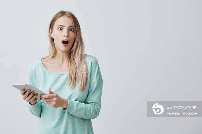 Isolated shot of shocked young blonde female wearing light blue sweater, looking and screaming with 