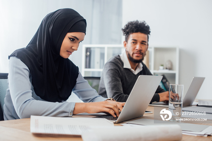 Selective focus of arabic businesswoman using laptop near african american colleague in office