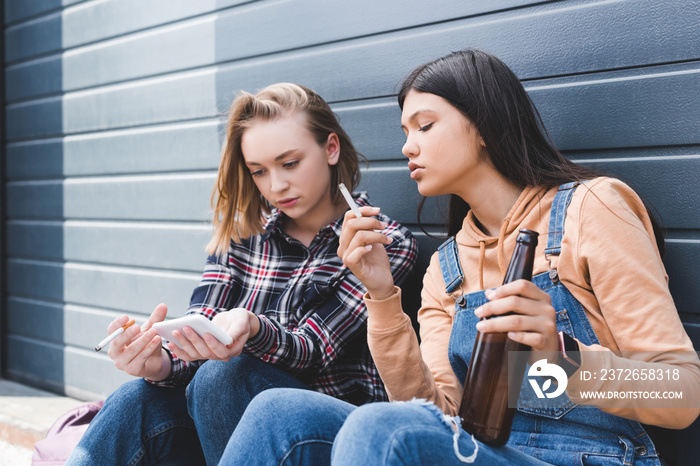 pretty friends smoking cigarettes, holding beer, sitting and looking at smartphone