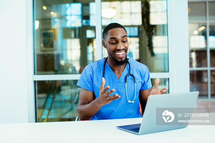 一位面带微笑的男医生的肖像，穿着蓝色磨砂服，使用笔记本电脑与患者在线交谈