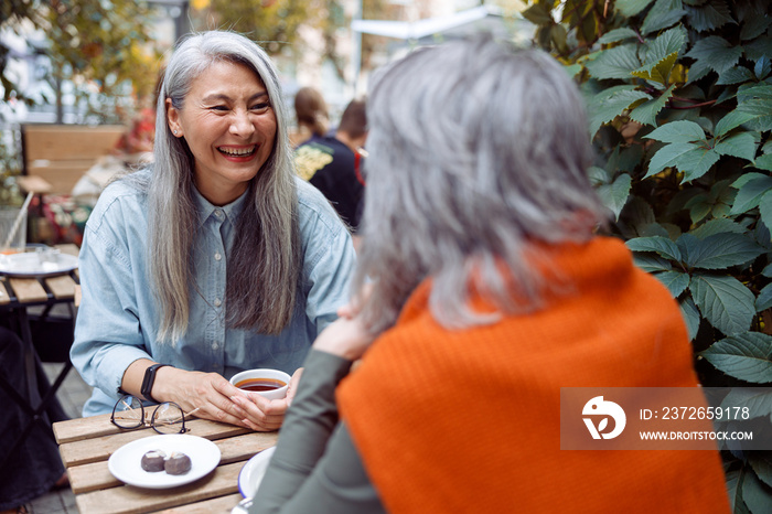 笑着的成熟亚洲女人和灰头发的朋友坐在户外咖啡馆的小桌子旁开会